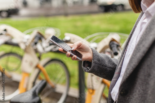 Close-up shot of hands using smartphone, young girl using bicycle rent mobile app smiling outdoors, female manager browsing smartphone standing near bike sharing, healthy city lifestyle