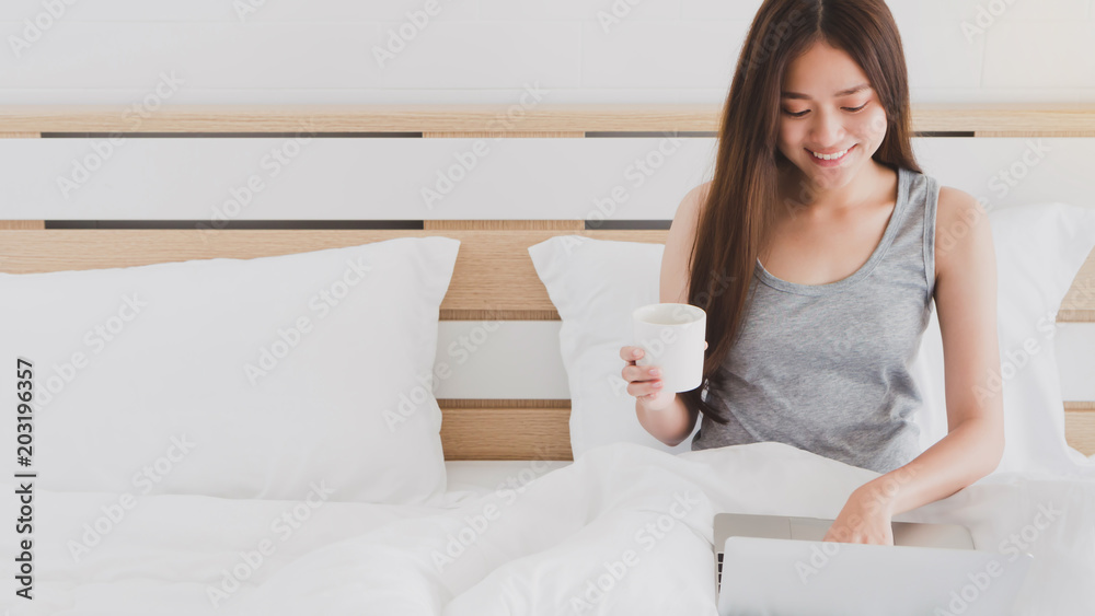 Asian beautiful smiling women using laptop while drinking coffee cup on white bed in bedroom interior morning.Concept of people use technology. Asian beautiful smiling women using laptop while drinking coffee cup on white bed in bedroom interior morning.Concept of people use technology.