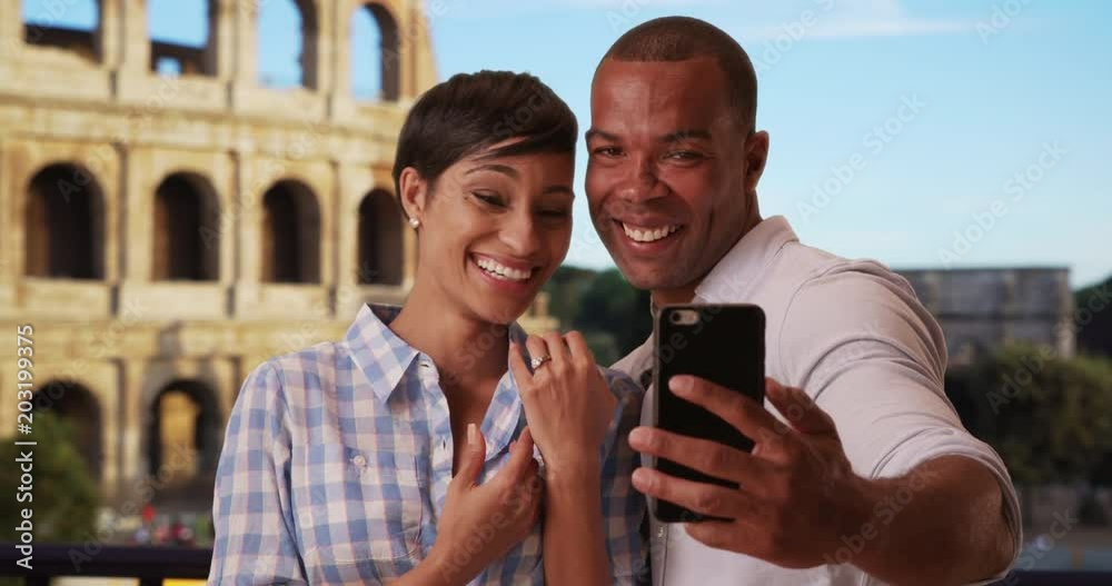 Newly engaged African male and female take photos together on a smartphone on their vacation in Rome, Italy, Overjoyed ethnic couple relishes their engagement near Roman Colosseum, 4k Newly engaged African male and female take photos together on a smartphone on their vacation in Rome, Italy, Overjoyed ethnic couple relishes their engagement near Roman Colosseum, 4k