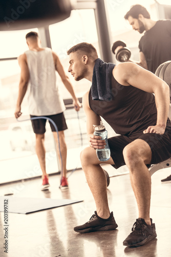 side view of handsome sportsmen resting and holding bottle of water in gym