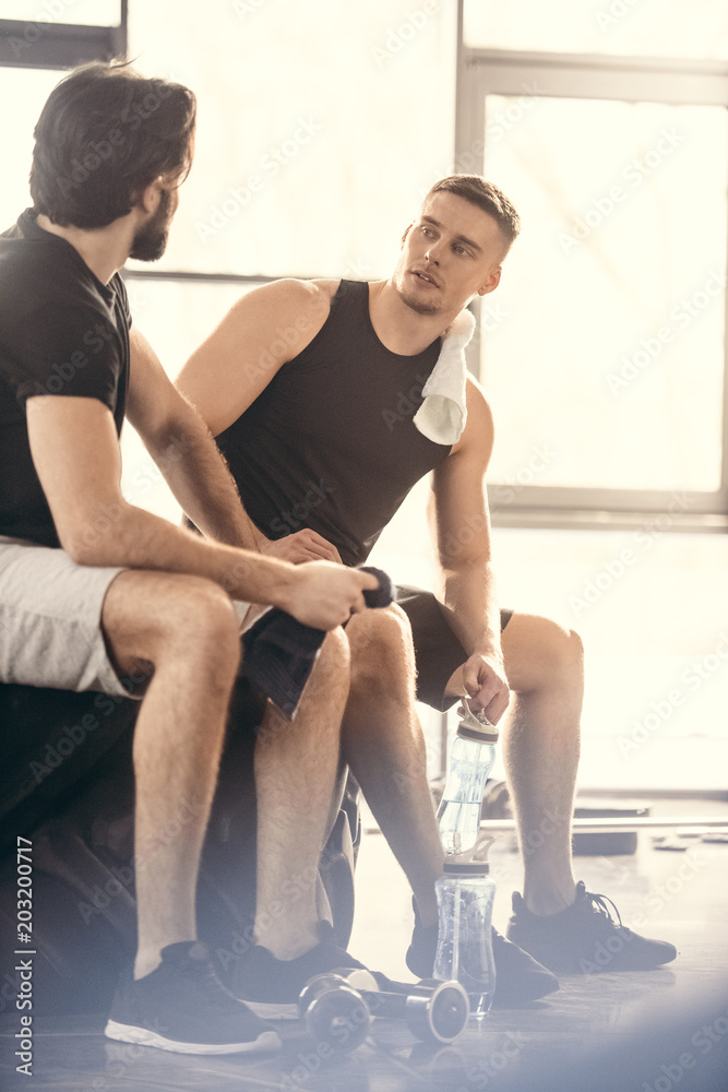 two young sportsmen sitting on tire and looking at each other after workout in gym