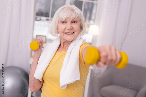 Gaining strength. Upbeat elderly woman with a towel around her neck exercising with a pair of dumbbells and smiling