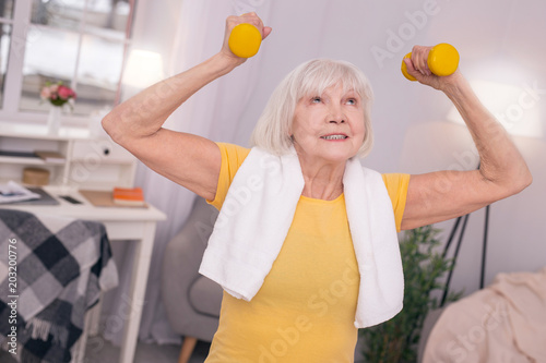 Exercising strength. Pleasant elderly woman with a towel around her neck lifting up dumbbells while working out in the living room in the morning