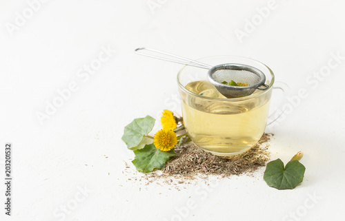 Cup with infusion of coltsfoot and flowers with leaves on white table.