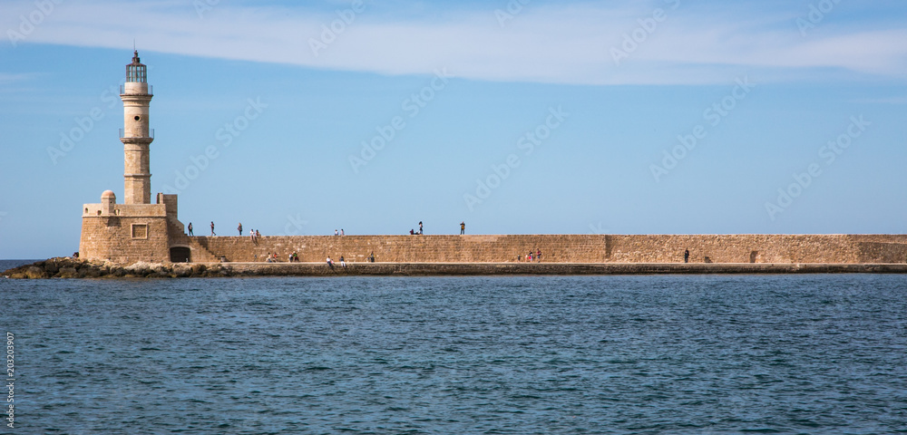 Leuchtturm am Ende einer lang gestreckten Kaimauer mit dem Meer im Vordergrund und blauem Himmel.