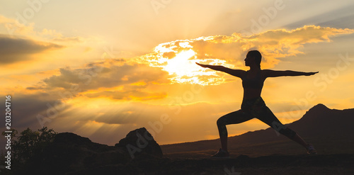 Silhouette of young woman practicing yoga or pilates at sunset or sunrise in beautiful mountain location, doing lunge exercise, standing in Warrior. © davit85