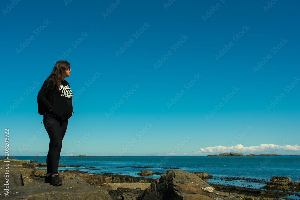 beautiful woman with dark hair dressed black standing on rock formations on coast and looking on ocean. 