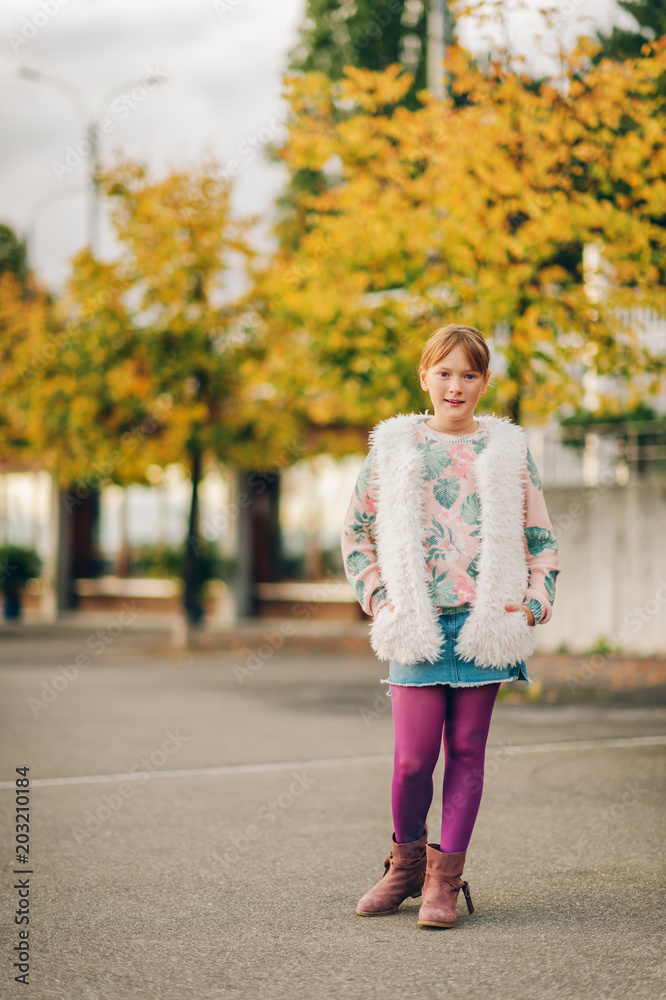 Outdoor fashion portrait of pretty kid girl wearing stylish pullover, white faux fur gilet and purple tights