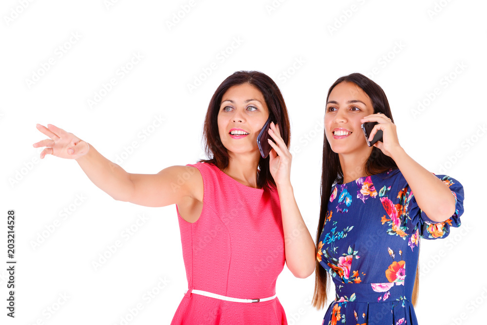 Two beautiful women, girlfriends, sisters in summer colored dress, pointing by finger and speak on mobile cell smartphone on a white background Two beautiful women, girlfriends, sisters in summer colored dress, pointing by finger and speak on mobile cell smartphone on a white background