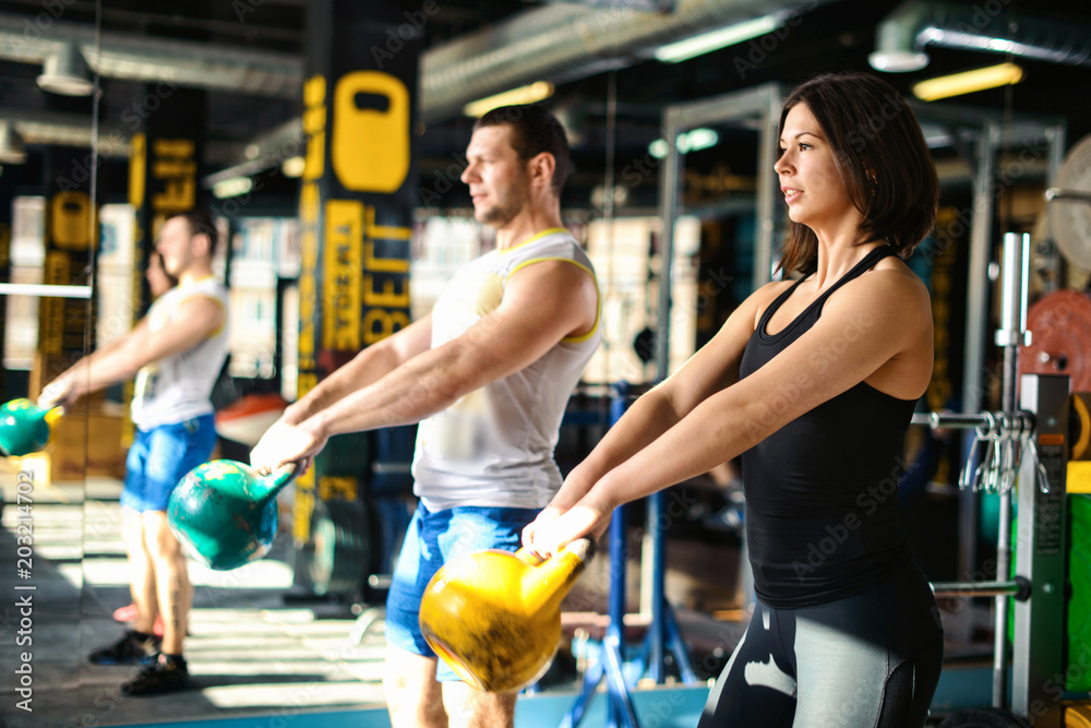 Close up front view of focused and motivated strong young fitness couple in sportswear doing exercizes crouching with the kettlebells reversed and doing squats in the gym at sunny day Close up front view of focused and motivated strong young fitness couple in sportswear doing exercizes crouching with the kettlebells reversed and doing squats in the gym at sunny day