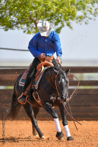 Widok z przodu jeźdźca w dżinsach, kowbojskich facetach i kraciastej koszuli na lejącym się koniu galopującym w czerwonej glinie na arenie.