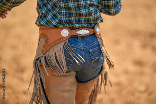 The rear view of cowgirl in jeans, cowboy chaps and checkered shirt in a ranch, on the red clay.  © PROMA