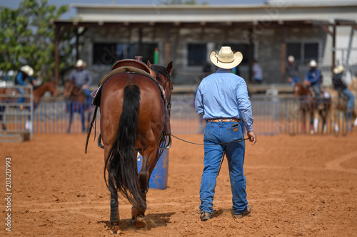 Cowboy in hat, jeans and white shirt accompanies his horse in a ranch, on the red clay. 