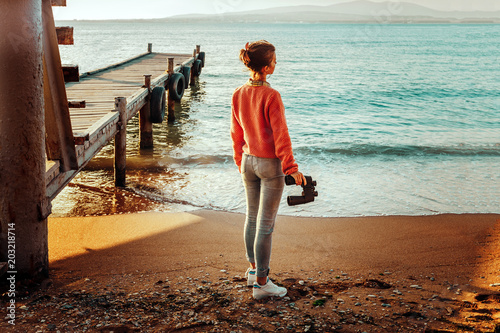 Fototapeta Naklejka Na Ścianę i Meble -  Girl Traveler With Binoculars In Hand Standing By The Sea Near Pier And Enjoying View Of Nature, Rear View