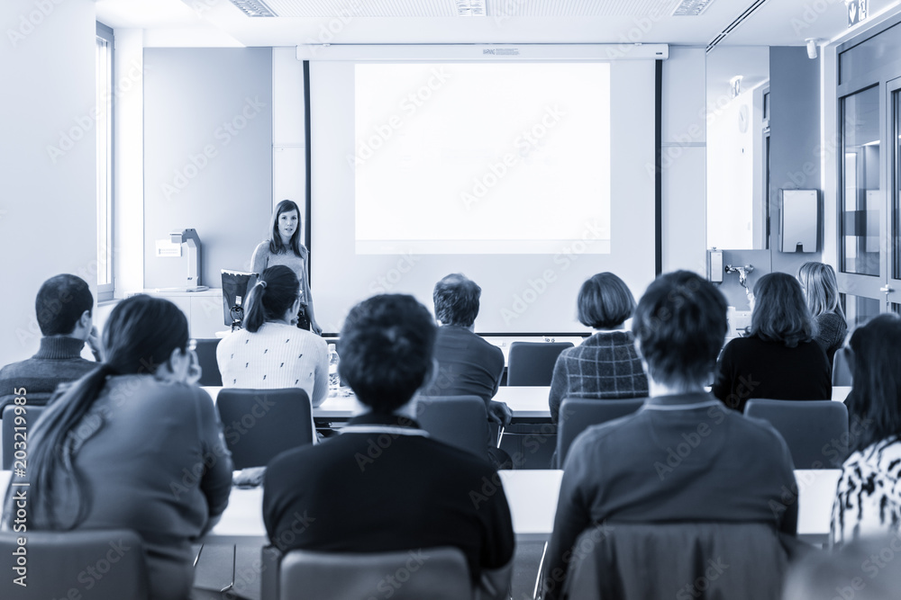 Female speaker giving presentation in lecture hall at university workshop.  Rear view of unrecognized participant in audience. Scientific conference  event. Black and white blue toned image. Photos | Adobe Stock