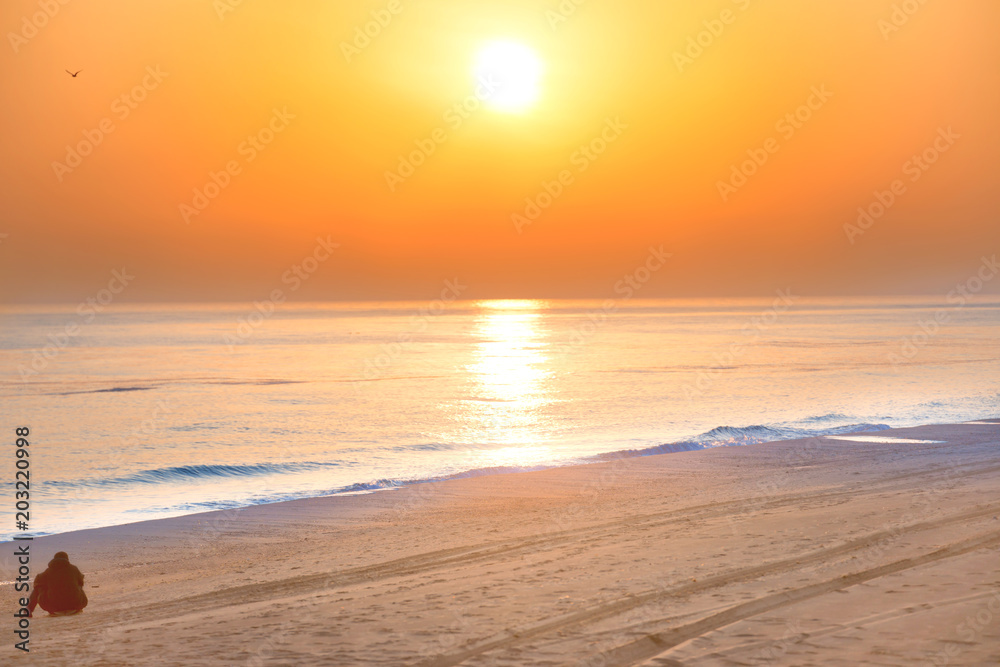 Sunset on the beach with man sitting on sand, long coastline, sun and dramatic sky 