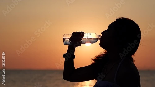 Silhouette of Asian woman drinking fresh water on tropical beach at sunset. Pregnant woman resting and drinking water outdoor in summer time.