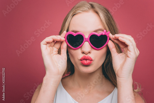 Adorable girl in glasses with pink edge is looking straight forward. he is holding glasses on its edge close to the head. Yng woman is making a kiss shape from her lips. Isolated on pink background.