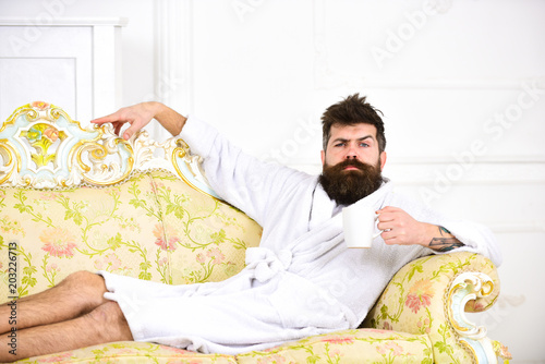 Man with beard and mustache enjoy morning while sitting on old fashioned luxury sofa. Man on thoughtful face in bathrobe drink coffee in luxury hotel in morning, white background. Luxury life concept. photo