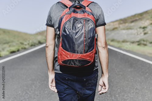 closeup of a young caucasian man, seen from behind, carrying a backpack walking by a secondary road