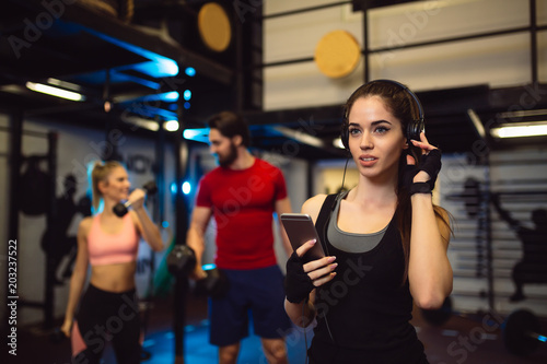 Young brunette with headphones listens music on her smartphone while her friends doing exercise in a gym