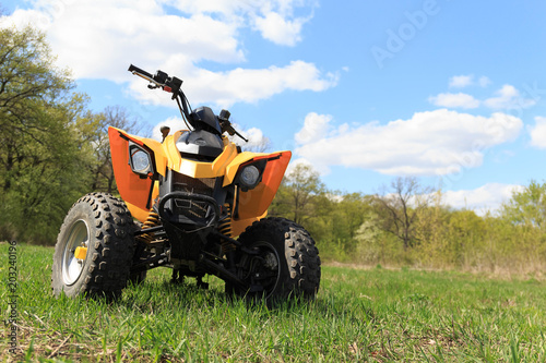 A four-wheeled yellow ATV quad-bike standing idle on the green grass, with trees and a blue sky with clouds on the background