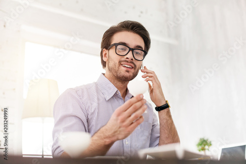 Wonderful news. Cheerful dark-haired man talking on the phone and holding a white object