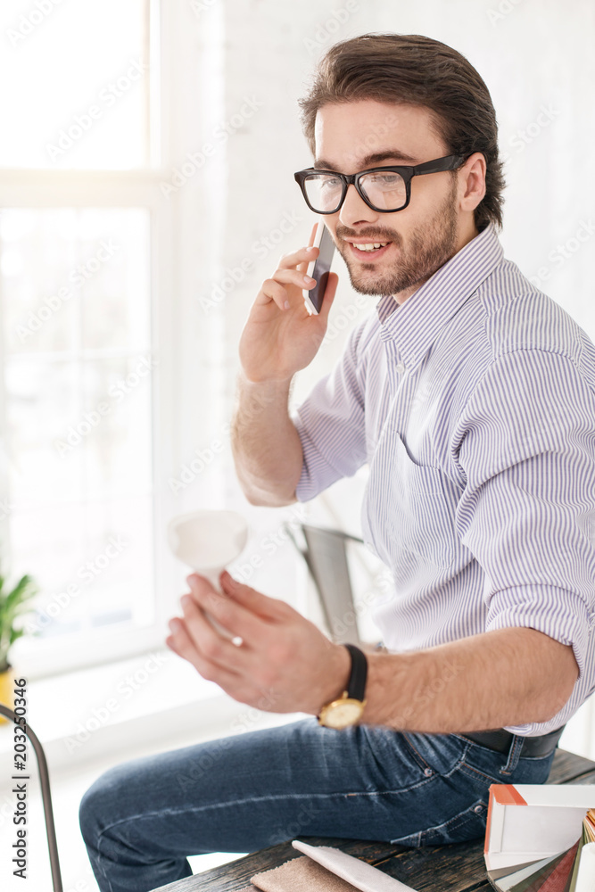 I am happy. Cheerful bearded man talking on the phone while sitting on the table