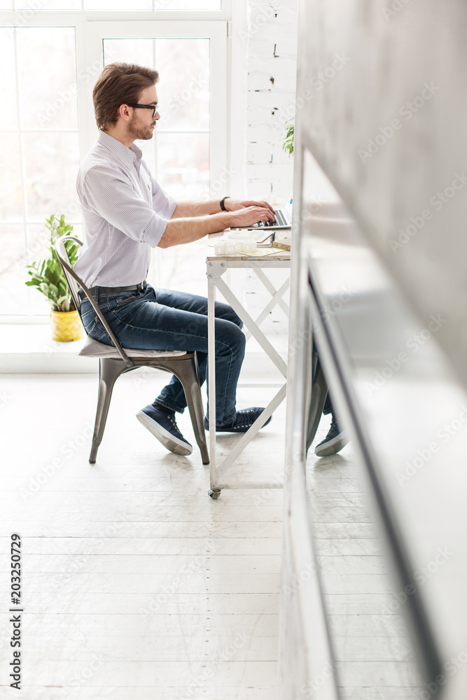 Working day. Serious bearded man sitting at the table and working on his laptop Working day. Serious bearded man sitting at the table and working on his laptop
