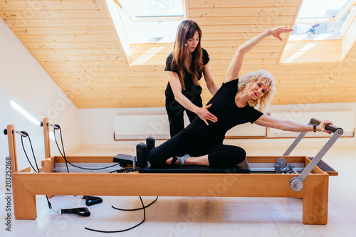 Sporty blond curly senior woman doing pilates exercises in gym with help from female physical therapist or instructor. photo