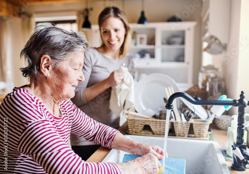 An elderly grandmother with an adult granddaughter at home, washing the dishes.