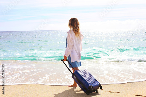 Young woman with her suitcase luggage on beautiful sandy beach w/ sea view on sunny day. Hipster female in loose cotton shirt on ocean shore having fun watching waves, sunbathing. Background, close up