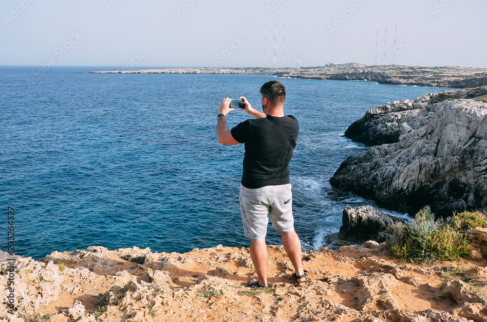 Man traveler taking photo with mobile phone standing near sea. Hiking man standing on top of mountain in Cape Greco, Cyprus, Mediterranean Sea. Travel, vacation concept. Copyplace, place for text