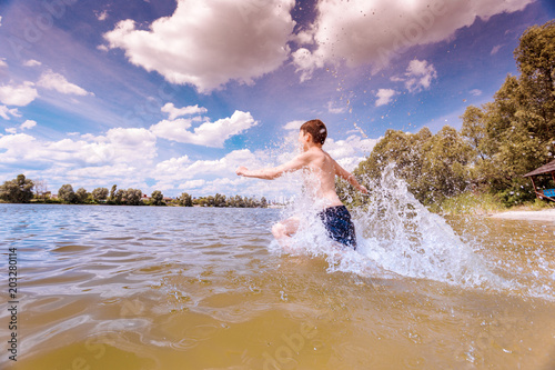 Happy boy running into the the lake water with splashes. Kid having fun outdoors. City outskirts.