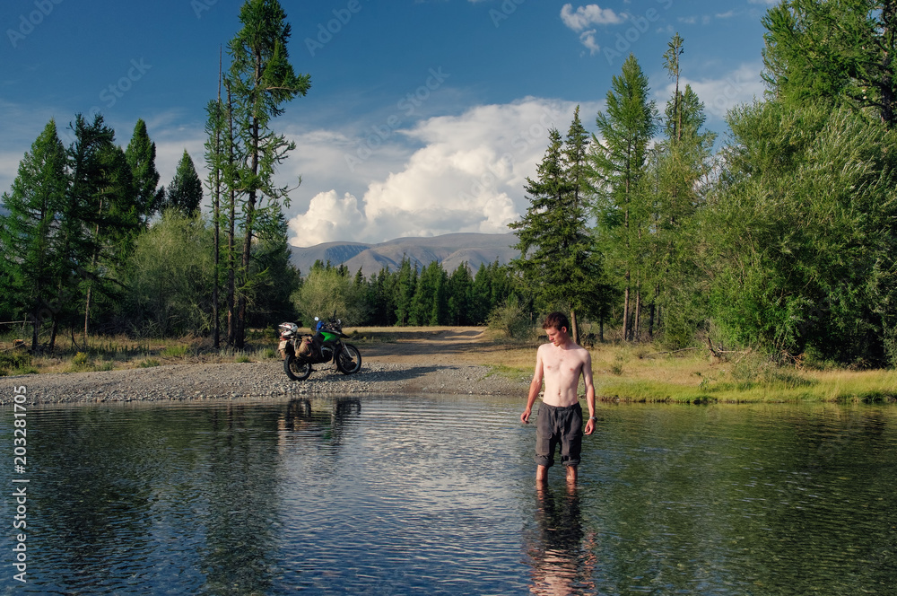 Man with nude body standing in water of mountain lake stream on the background of motorcycle bike among trees and highland steppe