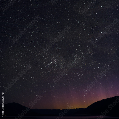 Night picture of a minimal landscape of the stars and Southern Cross in New Zealand. The purple, yellow, red and orange lights emerging behind the mountains came from Queenstown.