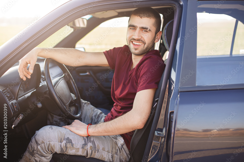 Happy brunet male drives his new car, sits behind wheel, has positive expression, enjoys long distance driving and high speed. Man driver sits in transport, looks directly into camera. Leisure, people Happy brunet male drives his new car, sits behind wheel, has positive expression, enjoys long distance driving and high speed. Man driver sits in transport, looks directly into camera. Leisure, people