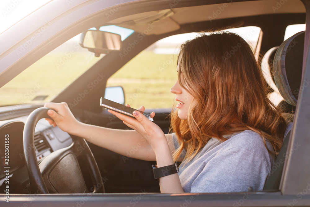 Sideways shot of concentrated female turns around wheel in car, makes voice call via cell phone, speaks with friends while drives automobile, does two things simultaneously. Technology and transport