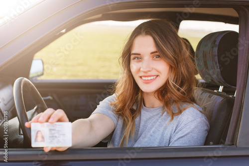 Obraz na płótnie Smiling young female with pleasant appearance shows proudly her drivers license, sits in new car, being young inexperienced driver, looks with joyful expression