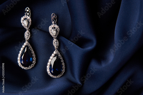 Two Golden sapphire earrings with small diamonds. Pair of platinum earring with sapphire gemstone on blue satin background. Luxury female jewelry, close-up