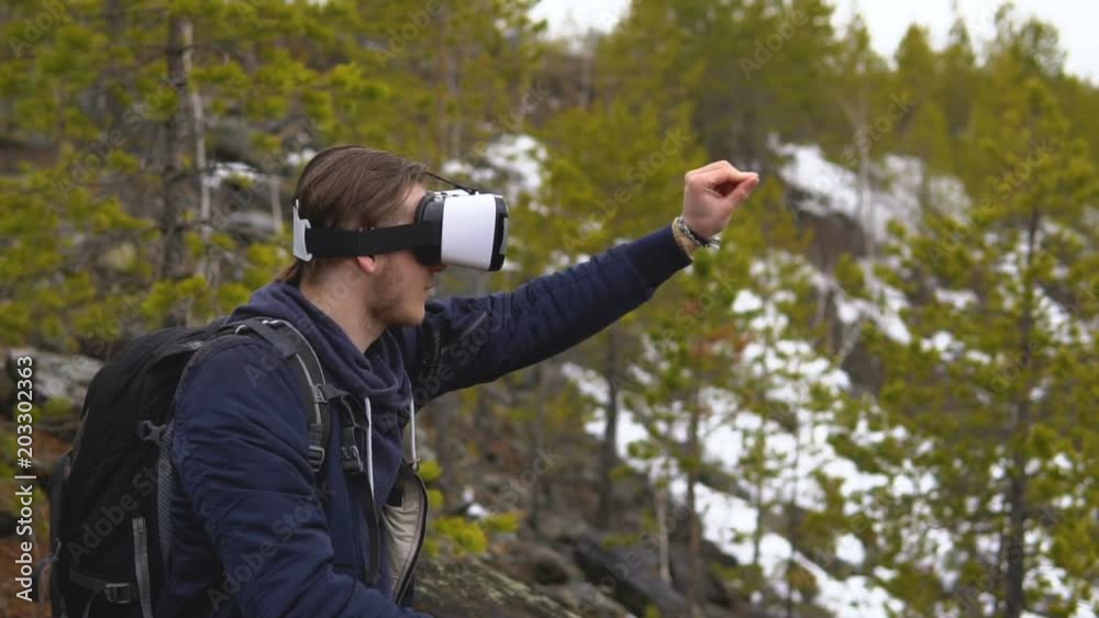 The artist-tourist in glasses of virtual reality draws a picture. In the background there is a coniferous forest and snowy mountain slopes. The artist-tourist in glasses of virtual reality draws a picture. In the background there is a coniferous forest and snowy mountain slopes.