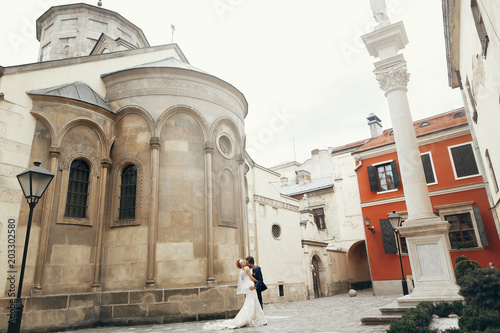 Handsome groom in blue suit sensually kissing gorgeous sexy bride in luxury white wedding dress outdoors, newlywed couple on a walk outdoors