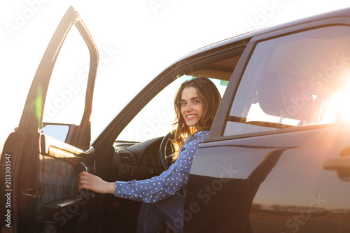 Positive woman sits in black car, looks with happy smile on face, tries to close door, ready for long trip, enjoys driving. Satisfied female driver glad to buy new transport. People, road, journey photo