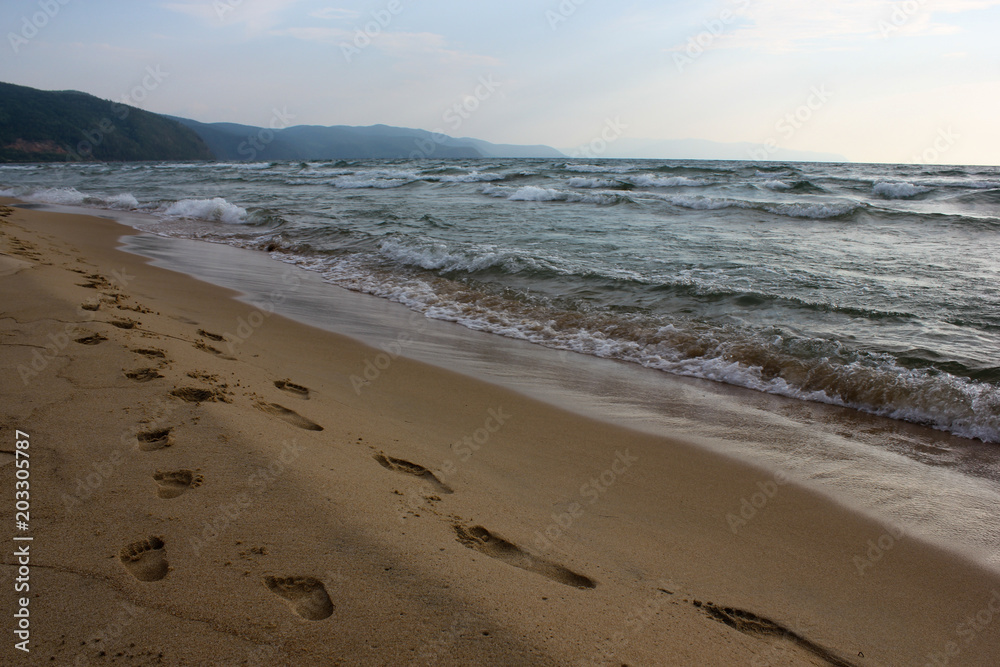 Close-up long path of family footprints on sandy ocean beach. Fathers or mothers and little sons steps in the sand of lake Baikal seashore near the water and mountain in the evening sunset Close-up long path of family footprints on sandy ocean beach. Fathers or mothers and little sons steps in the sand of lake Baikal seashore near the water and mountain in the evening sunset