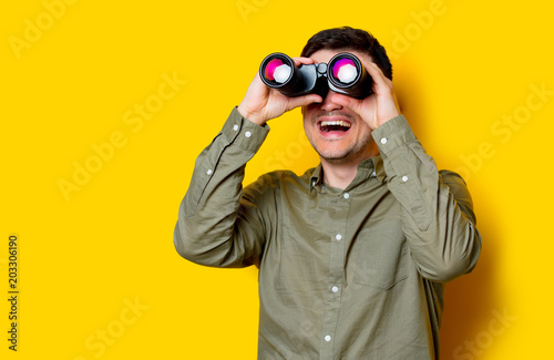 Billede på lærred Young man looking for something with binoculars on yellow background