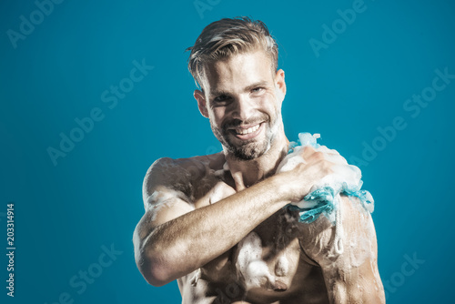 Handsome bearded macho, unshaven man with strong muscles taking shower. Man with naked torso is washed in shower. Muscular guy covered with foam. Smiling man washing body in bathroom. Copy space.