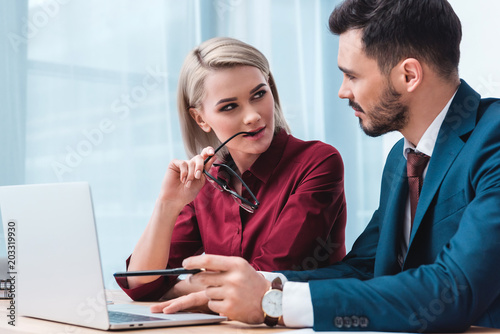 yiung businesswoman holding eyeglasses and flirting with handsome male colleague in office