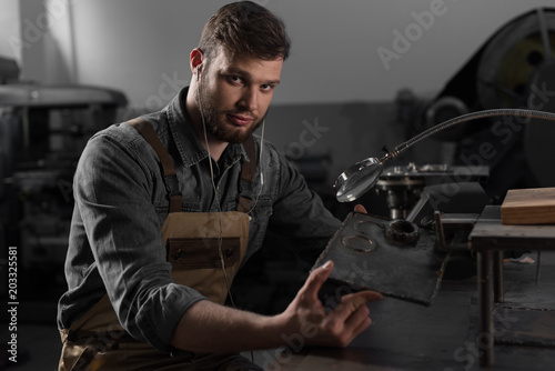 Portrait of young male worker in earphones holding metal part under magnifying glass
