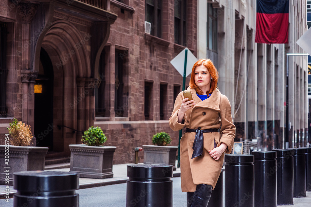 Young American businesswoman traveling in New York, wearing long brown woolen overcoat, sitting on metal pillar on narrow vintage street, texting on cell phone, looking up, thinking, taking work break