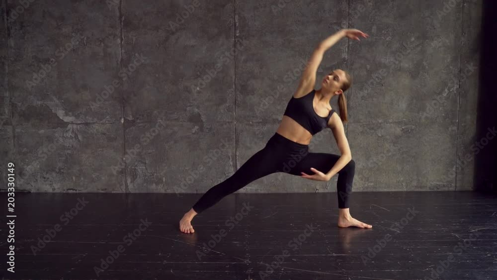 A beautiful, smart, athletic girl does yoga exercises in a gym in loft style, with natural light from large windows. She is dressed in a sporty uniform, breeches and crocheted top. Stretching. 4 k
